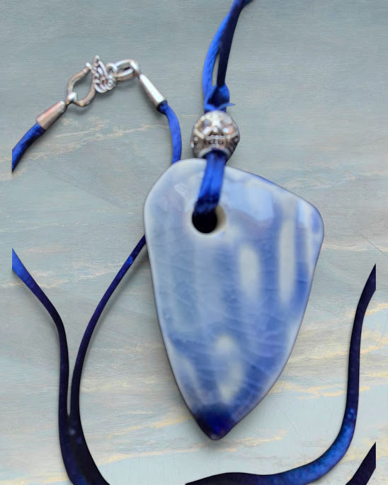 Heartfelt Creations artisan a unique, handcrafted ceramic pendant with a crackled white glaze and a deep blue base, featuring a natural, rough-cut crystal embedded in its center, suspended from a vibrant blue silk cord with a decorative silver-toned bead --backside