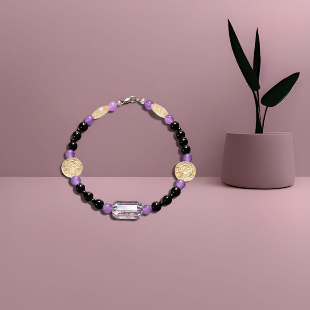 A beaded bracelet with black and purple beads, gold coin charms, and a clear rectangular faceted bead, displayed on a pink surface next to a potted plant.