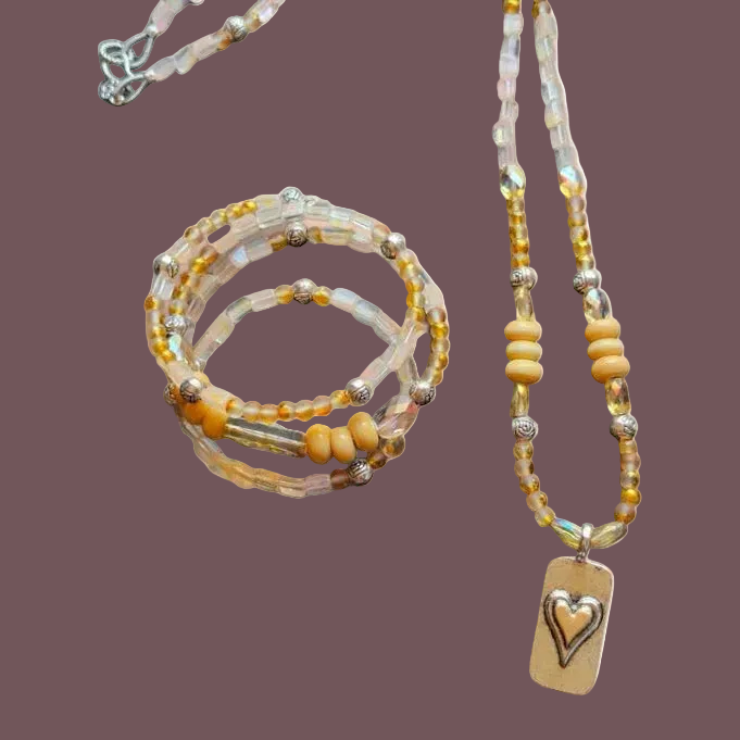 A beaded jewelry set featuring a long necklace with a rectangular heart-shaped pendant and a stack of three beaded bracelets, displayed on a solid mauve background.