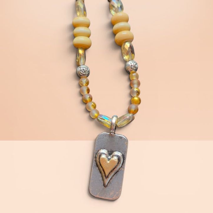 A beaded necklace featuring pale yellow and iridescent beads with a silver-toned rectangular pendant that has a raised gold heart design, displayed on a split beige background.