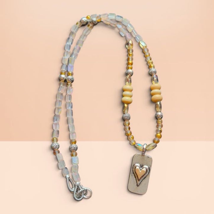 A beaded necklace with iridescent pale blue beads, yellow and gold spacer beads, a silver-toned rectangular pendant, and a heart-shaped inlay, displayed on a peach-colored background.