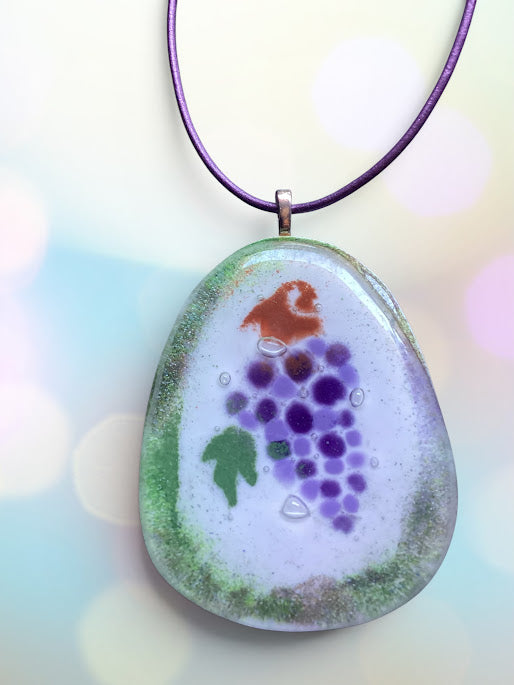 A handmade glass pendant necklace featuring a grapevine design with purple grapes, a green leaf, and a small orange bird on a white background, suspended from a purple cord.