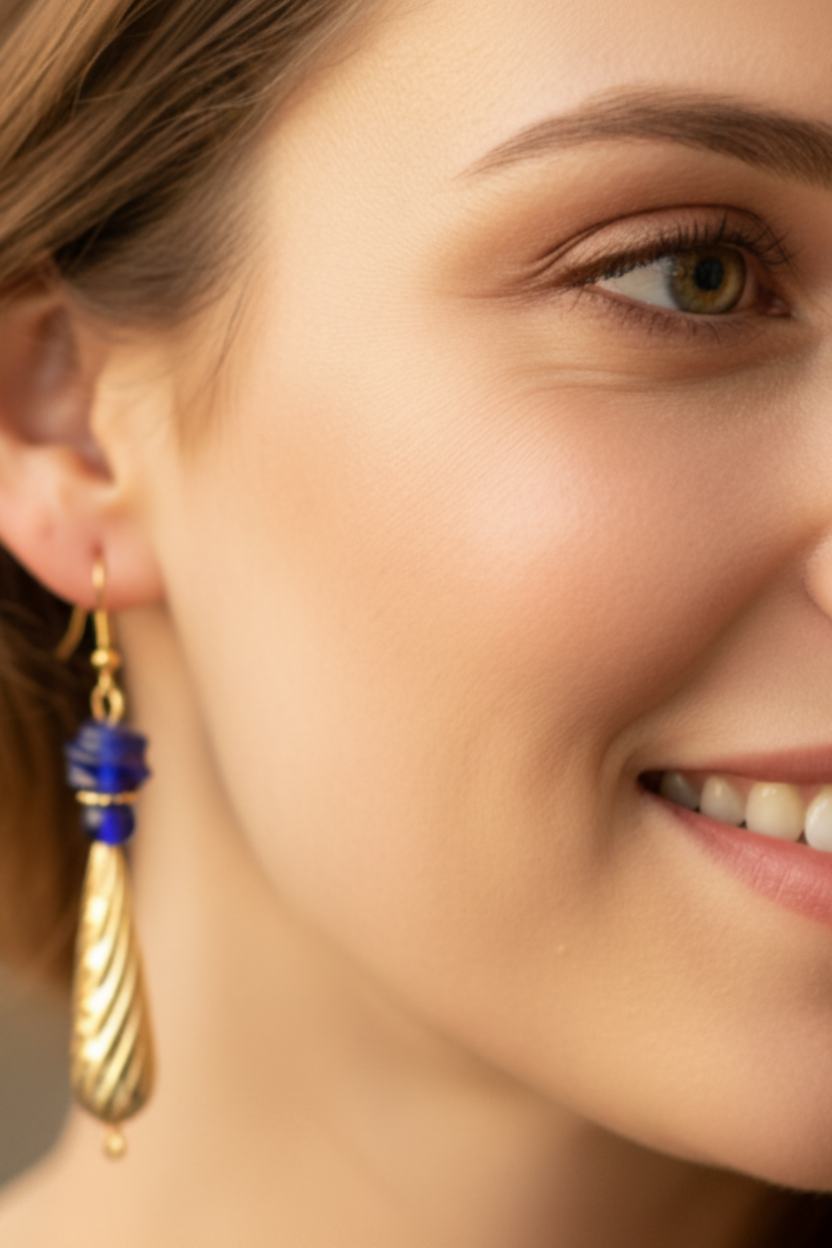 A close-up of a woman wearing a gold teardrop-shaped dangle earring with blue bead accents, shown against a softly blurred warm-toned background.