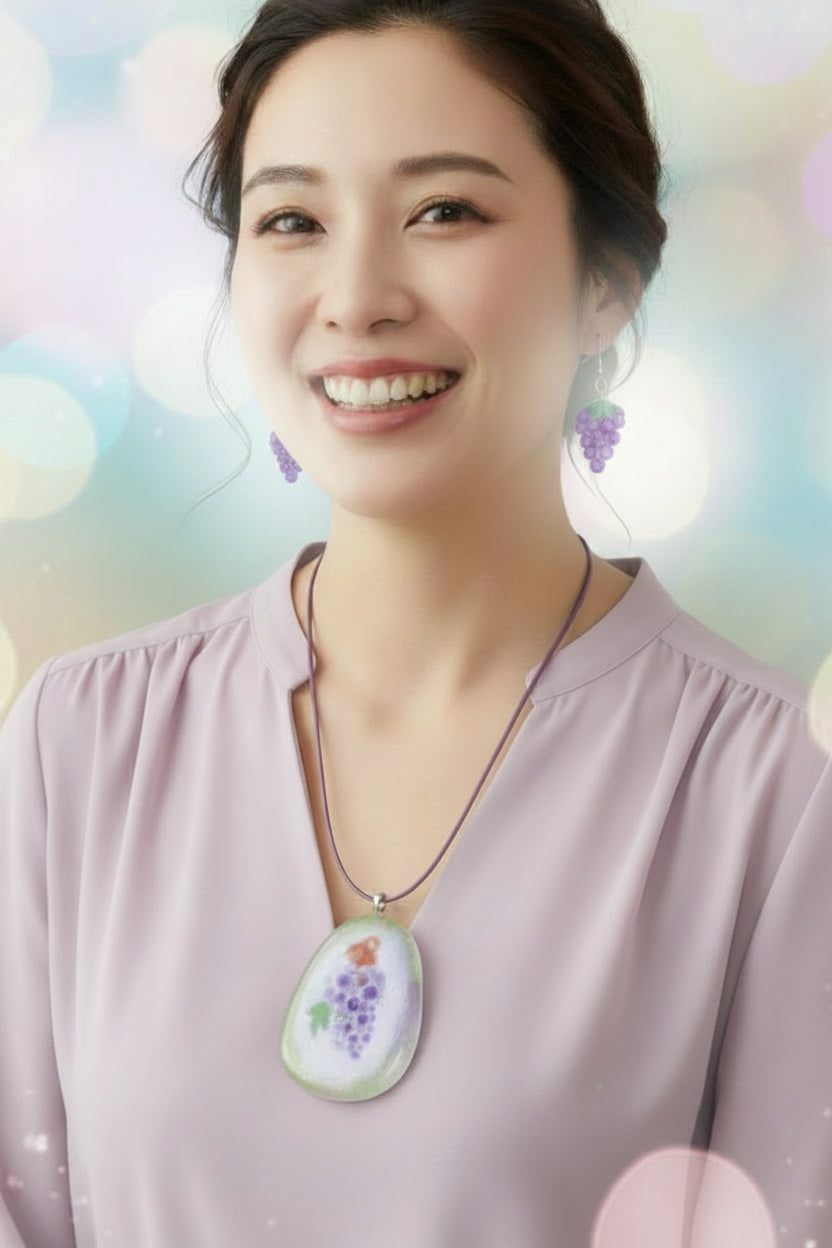 A glass pendant necklace and matching grape-shaped drop earrings on a soft pastel background. The pendant has a purple cord and a clear glass body with a painted purple grape cluster, green leaves, and a small orange detail.