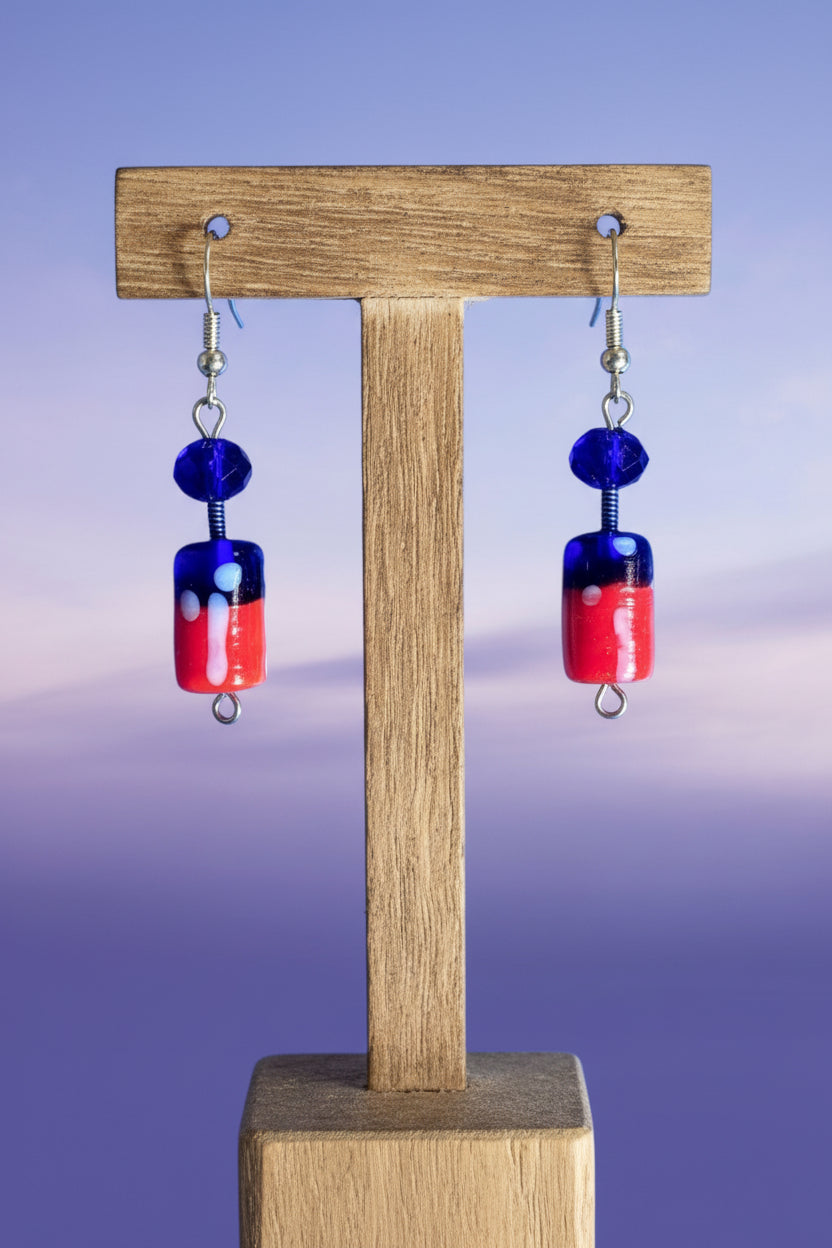 A pair of handmade glass dangle earrings with silver fishhook wires. Each earring features a faceted dark blue bead, a wrapped blue-and-red glass cylinder bead with white swirls, and a small round blue glass bead below.