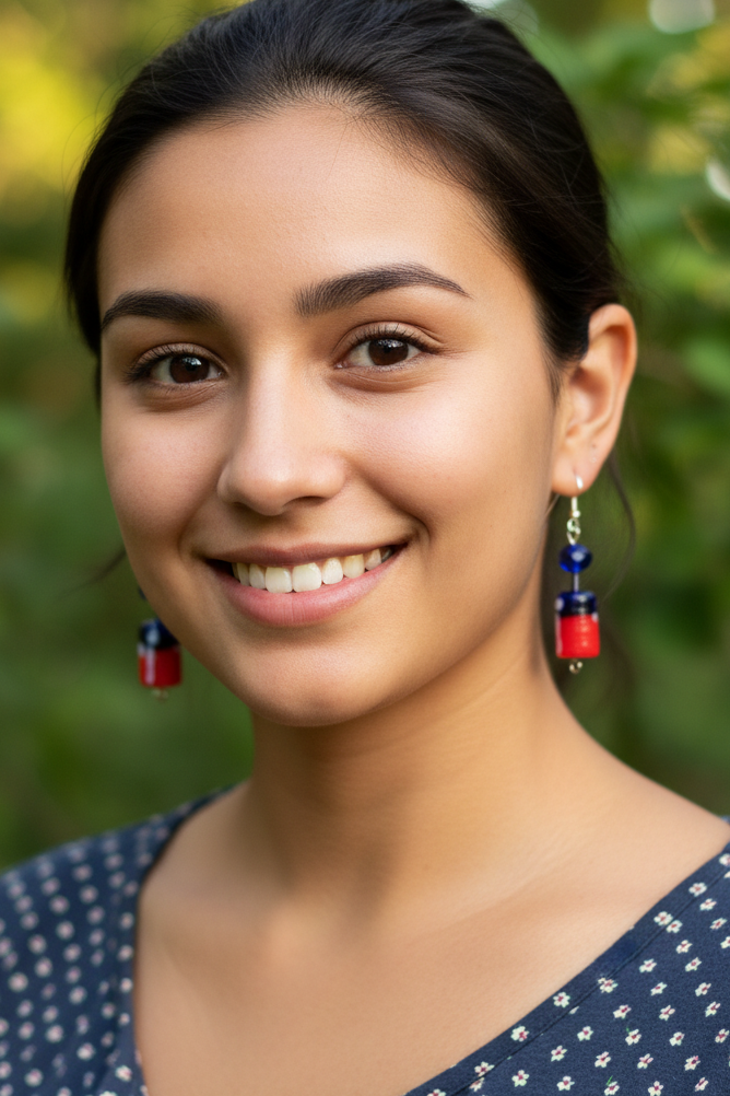 A smiling woman with dark hair pulled back, wearing dangling earrings with red and blue beads and a navy blouse with small white floral prints, set against a blurred green outdoor background.