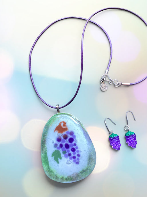 A handmade glass pendant necklace with a purple cord and a grapevine design, shown alongside matching grape-shaped dangle earrings on a pastel bokeh background.