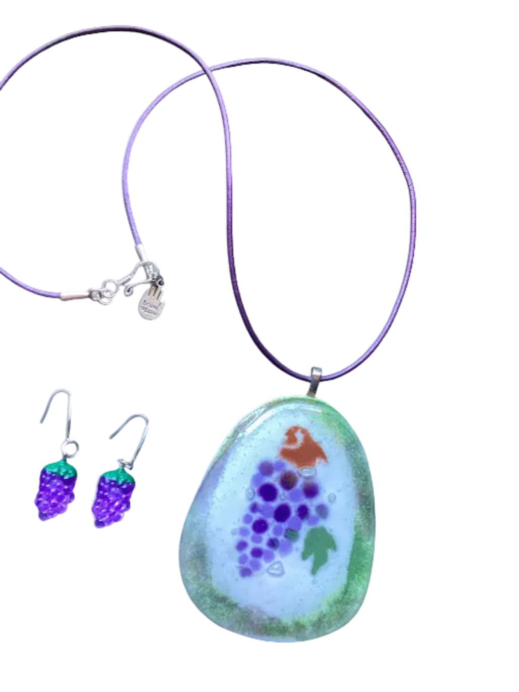 A glass pendant necklace and matching grape-shaped dangle earrings on a plain white background. The necklace has a purple cord and a large irregular glass pendant with a white center, purple grape motif, green border, and small orange detail.