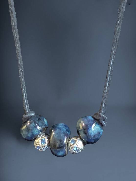 Heartfelt Creations'  handmade necklace featuring three large, glossy, deep blue home-made ceramic beads with a mottled texture, separated by two ornate, silver-toned metal spacer beads with a star-like pattern and small blue crystal accents, all strung on a dark cord.