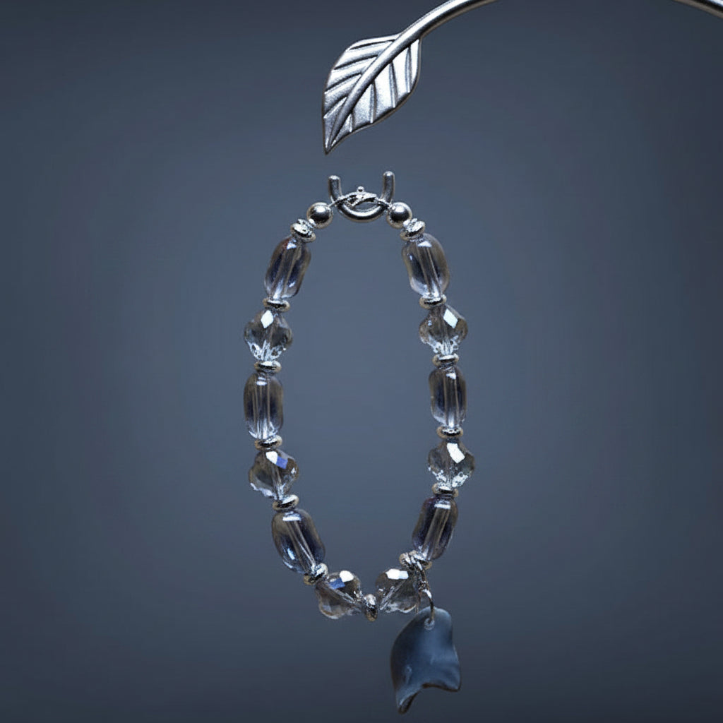 A beaded bracelet made of faceted clear glass beads with a small dark blue-gray shell charm, displayed on a dark gray gradient background.
