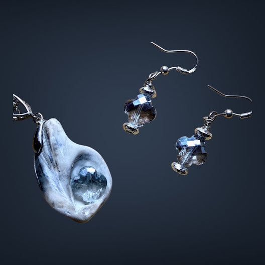 A jewelry set featuring a large irregular white and blue stone pendant and two matching dangling earrings with faceted blue glass beads and silver hooks, shown on a dark gradient background.