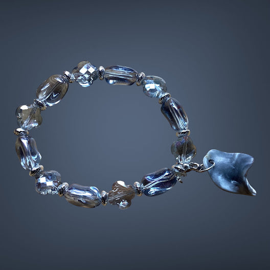A beaded bracelet made of faceted clear glass beads with a small dark blue-gray shell charm, displayed on a dark gray gradient background.