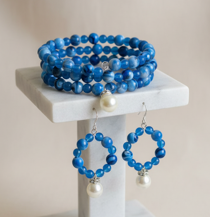 Blue beaded jewelry set including a multi-strand bracelet, hoop earrings, drop earrings, and a small beaded necklace, displayed on a white marble stand against a soft beige background.