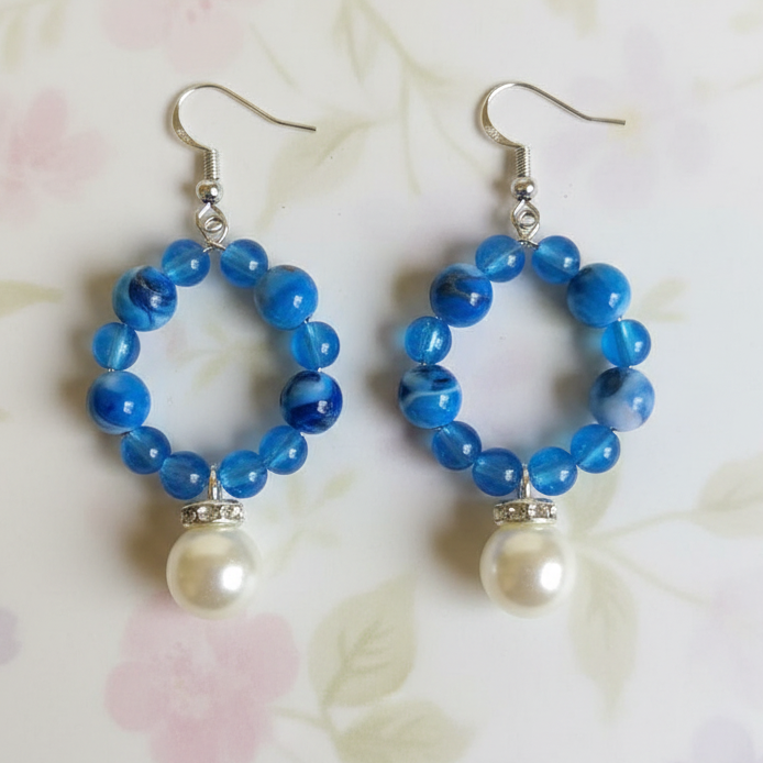 A pair of handmade dangle earrings featuring blue marbled beads in circular hoop shapes, each hanging from a silver fishhook ear wire with a small white pearl bead dangling below. The earrings are displayed on a light background with a soft pink and green floral pattern.