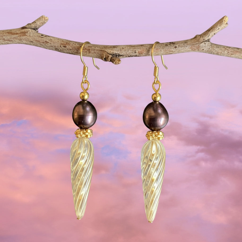 A pair of handmade dangle earrings featuring dark purple freshwater pearls, gold wire hooks, and textured gold glass drops, set against a pink and purple cloudy sky background.