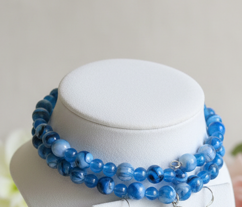 A blue beaded jewelry set displayed on a white jewelry bust, featuring a double-strand beaded necklace and matching beaded hoop earrings with white pearl-like drops, set against a soft beige background with blurred pink and white flowers.