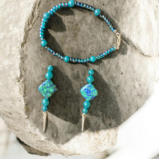 A jewelry set featuring a beaded bracelet and matching stud earrings with turquoise-blue beads and a central square opal-like stone, displayed on a textured stone surface.