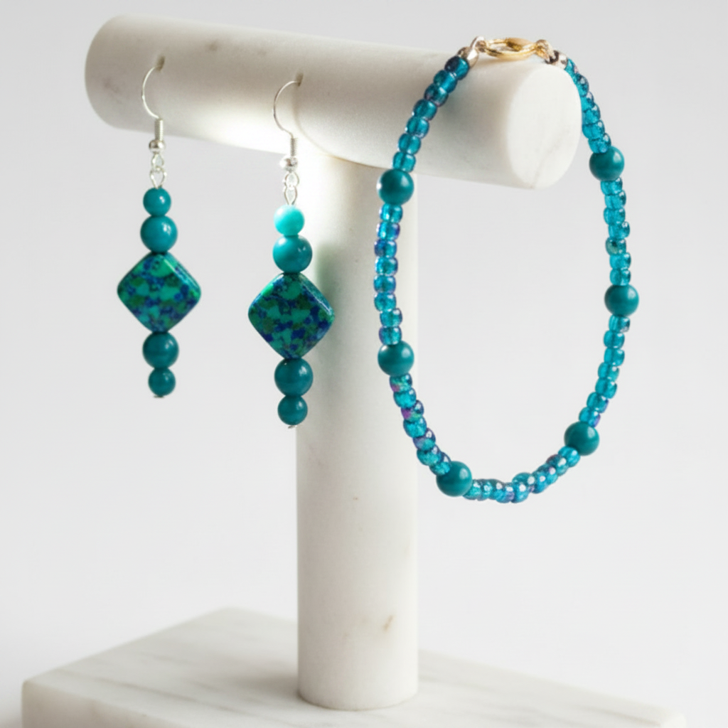 A beaded bracelet and matching drop earrings displayed on a white marble jewelry stand against a clean white background. The jewelry features teal and turquoise beads with a small gold clasp on the bracelet and silver hooks on the earrings.