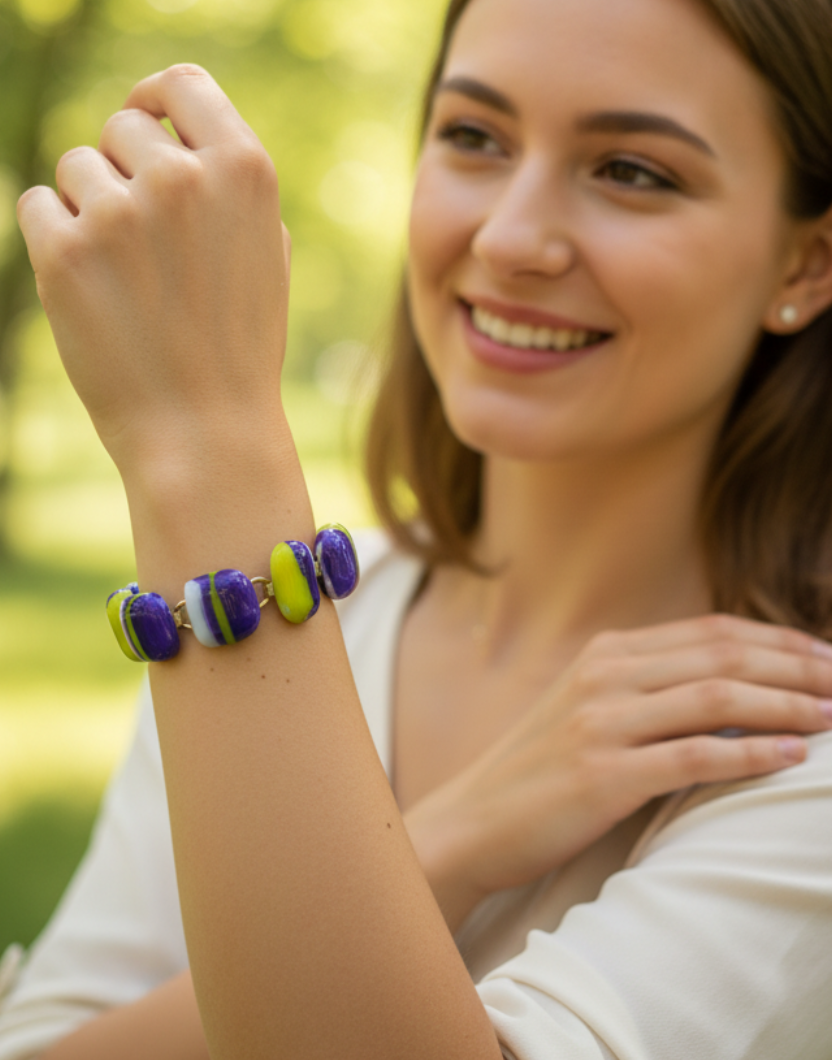 A woman outdoors wearing a colorful beaded bracelet, smiling and looking to the side. The bracelet features alternating purple, blue, and lime green beads.