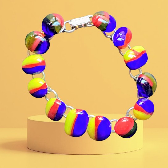 A colorful handmade ceramic glass bead chain bracelet with marbled segments of blue, yellow, red, and black, displayed on a light beige pedestal against a solid warm yellow background.