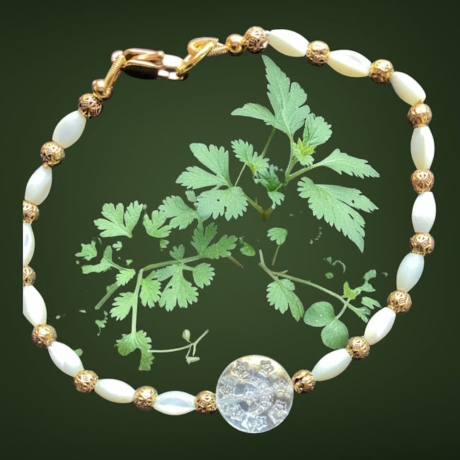 A beaded bracelet featuring white iridescent oval beads, textured gold spacer beads, and a central clear faceted disc charm, displayed against a dark green background with a botanical illustration of green leaves.
