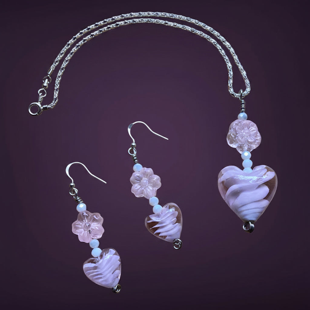 Heartfelt Creations offers a matching set of handcrafted jewelry, including a necklace and a pair of earrings, featuring silver-toned chains and hooks, with each piece adorned with a pale pink, swirled lampwork  glass heart bead and a delicate, clear glass flower bead.