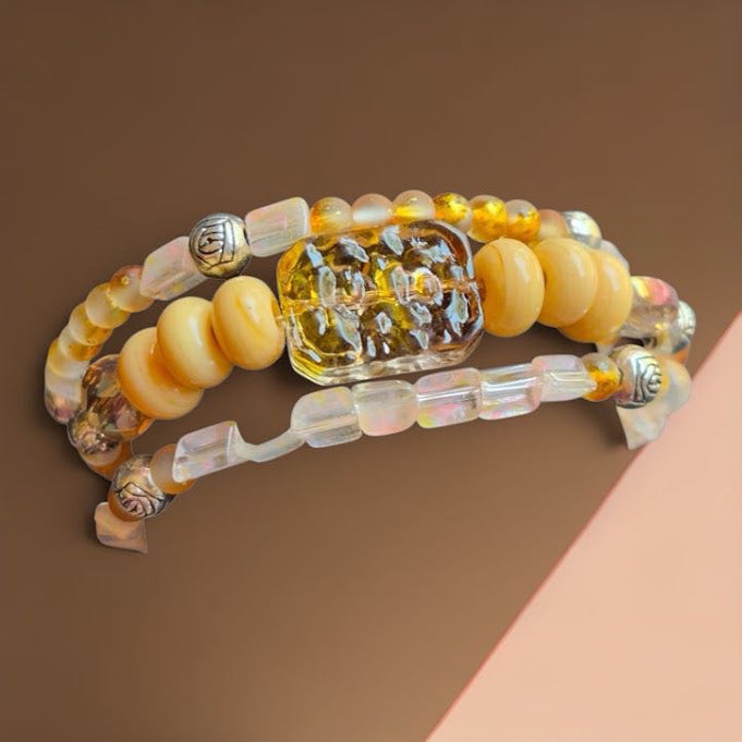 Three beaded bracelets arranged in a stack on a brown background with a peach-colored corner accent. The bracelets feature pale yellow round beads, translucent iridescent beads, a large faceted yellow rectangular centerpiece, and small silver-toned decorative beads.