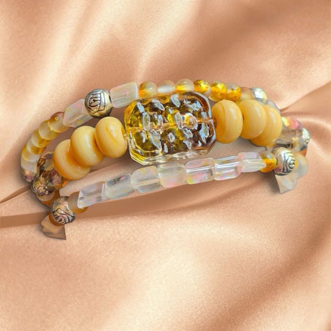 A set of three beaded bracelets with yellow and cream beads, clear faceted beads, and silver-toned metal charms, arranged on a draped peach satin fabric.