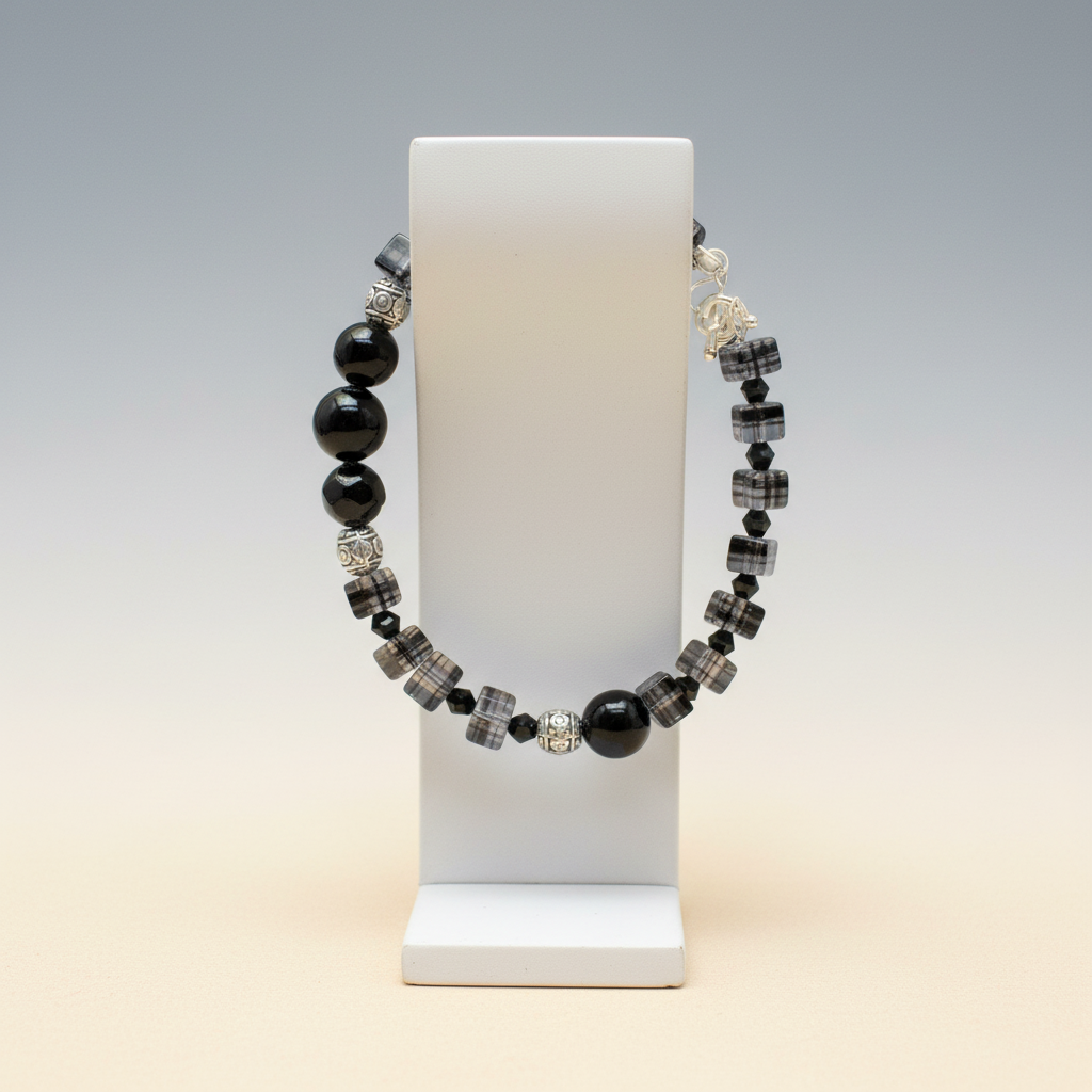 A beaded, clasped bracelet displayed on a white jewelry stand. The bracelet features glossy black beads, faceted black and gray striped beads, and small clear crystal accent beads, set against a soft gray-to-beige gradient background.