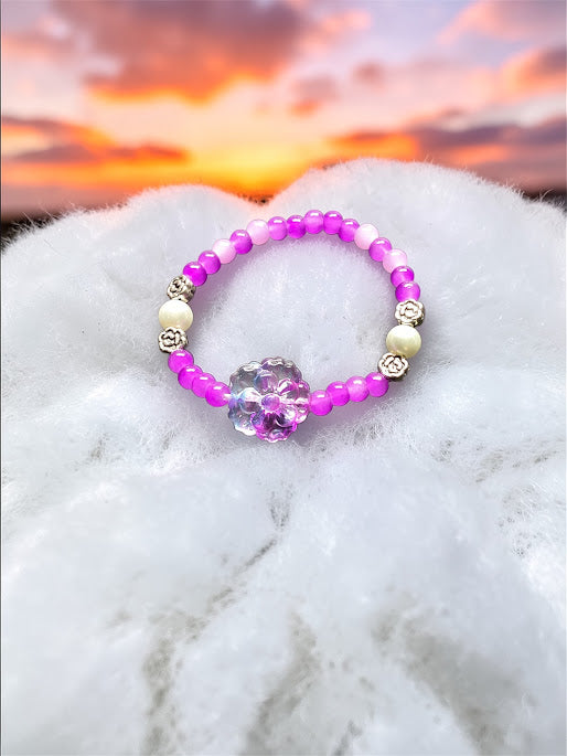 This delightful, hand crafted bracelet is a perfect way to add a pop to any outfit.   A beaded stretch bracelet features purple, lavendar and magenta beads, white pearl-like beads, silver-toned spacer details, and a central clear acrylic flower charm.  Simple yet Stunning!