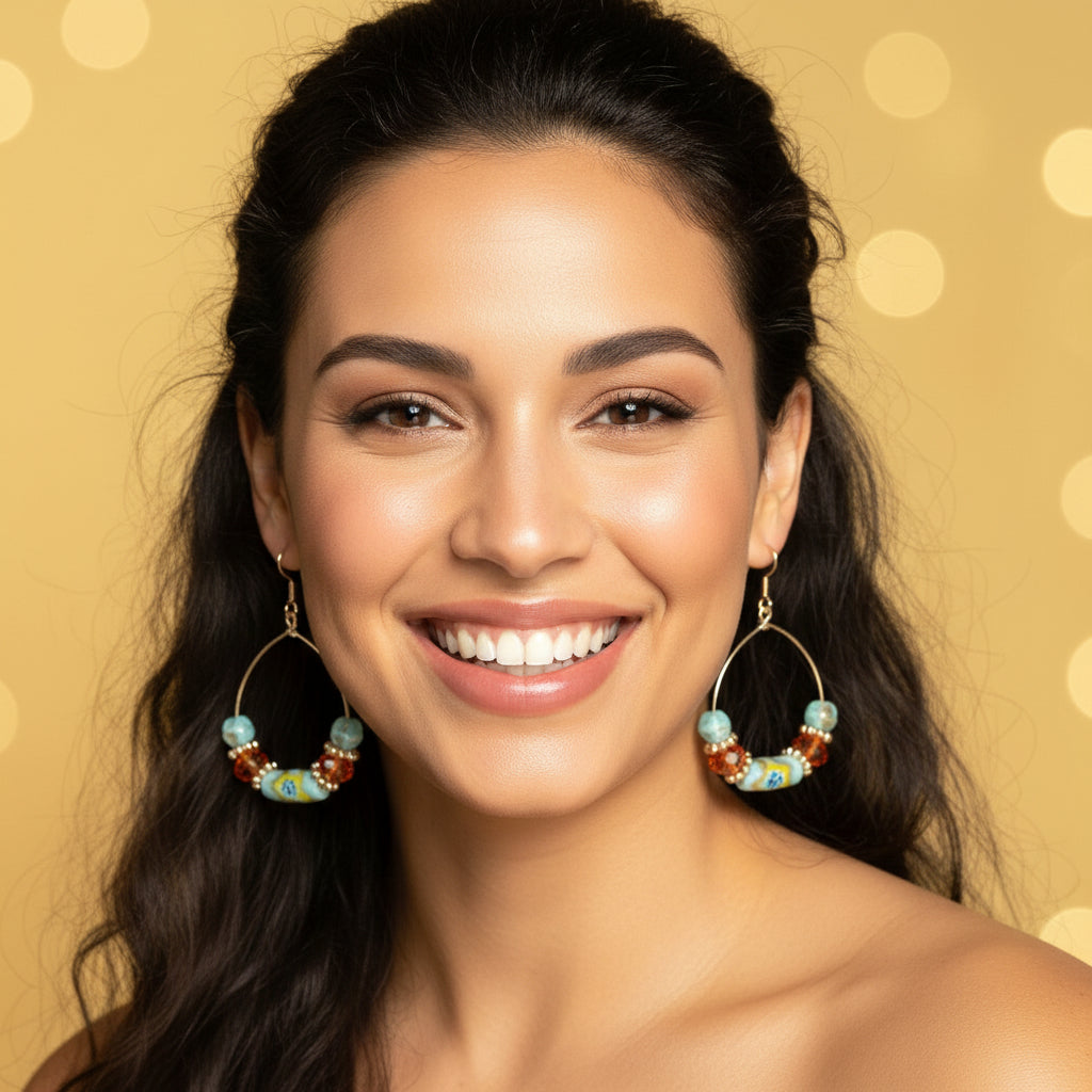 A pair of handmade hoop earrings with silver-tone metal hoops, light blue beads, orange faceted beads, and a central marbled bead section, hanging from a natural branch against a warm yellow bokeh background.