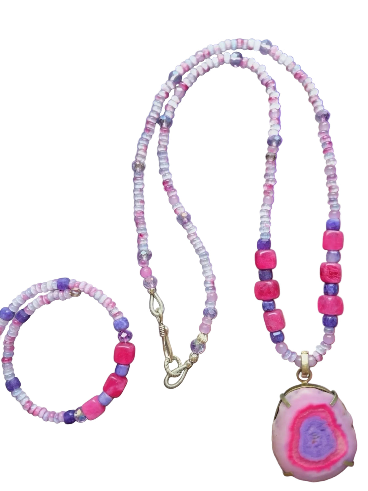 Heartfelt Creations handmade jewelry set consisting of a necklace and a bracelet, both featuring a mix of small, faceted beads in shades of white, pink, and light purple, interspersed with larger, square-shaped beads in vibrant magenta and deep purple. The necklace has a large, oval-shaped pendant with a polished, pink and purple agate slice set in a gold-toned bezel, and a silver-toned hook clasp. The bracelet is a smaller version of the necklace, with the same bead pattern.