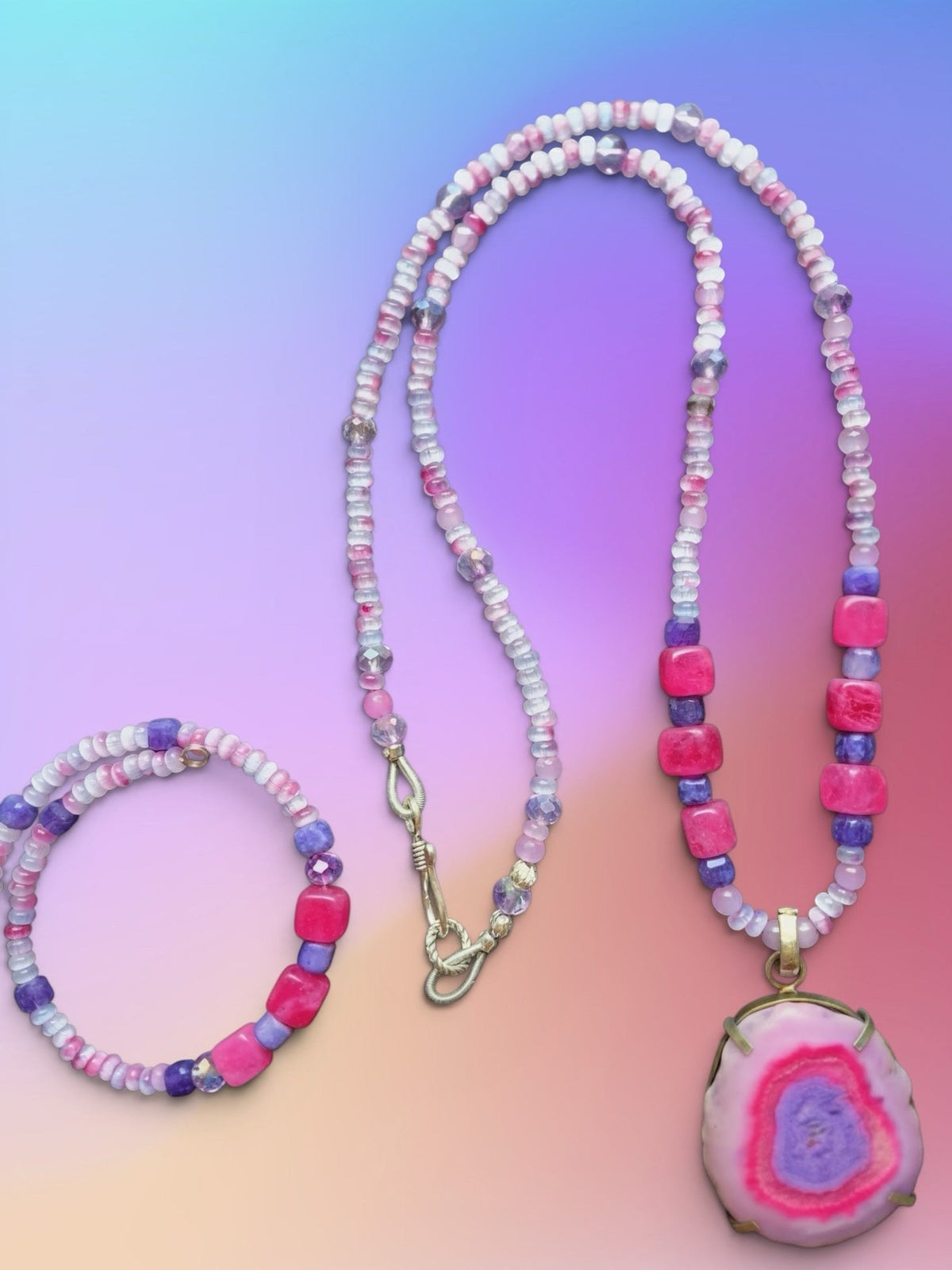 Heartfelt Creations handmade jewelry set consisting of a necklace and a bracelet, both featuring a mix of small, faceted beads in shades of white, pink, and light purple, interspersed with larger, square-shaped beads in vibrant magenta and deep purple. The necklace has a large, oval-shaped pendant with a polished, pink and purple agate slice set in a gold-toned bezel, and a silver-toned hook clasp. The bracelet is a smaller version of the necklace, with the same bead pattern.
