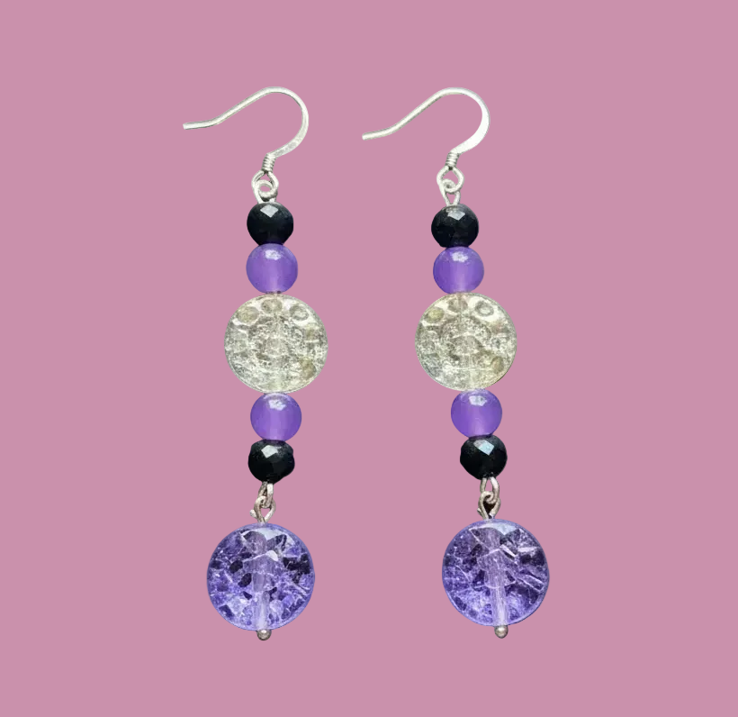 A pair of handmade dangle earrings featuring purple amethyst beads, black beads, a clear faceted round bead, and silver-tone hooks, displayed on a dark gray textured surface.