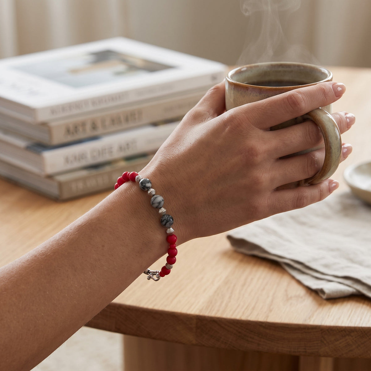 Heartfelt Creations beaded bracelet with red matte and grey stone beads worn on a wrist holding a steaming coffee mug