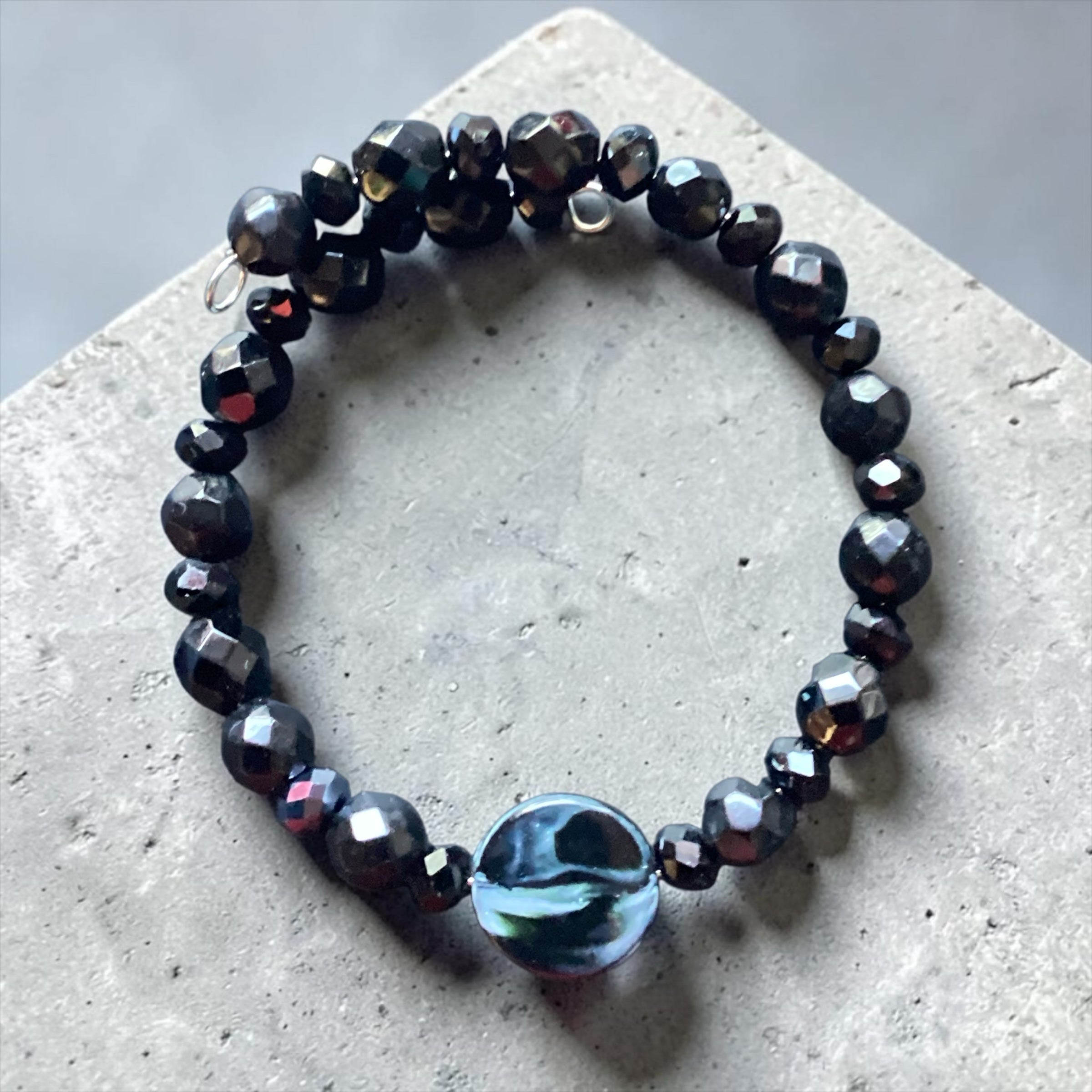 Heartfelt Creations black faceted beaded wrap bracelet with a central iridescent circular charm on concrete surface.