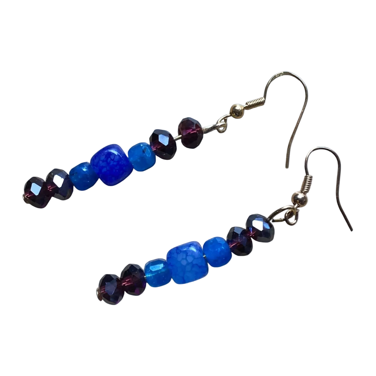 A pair of drop earrings with faceted beads in shades of royal blue, cobalt blue, and deep purple, suspended from silver-tone fishhook wires, shown on a plain white background.