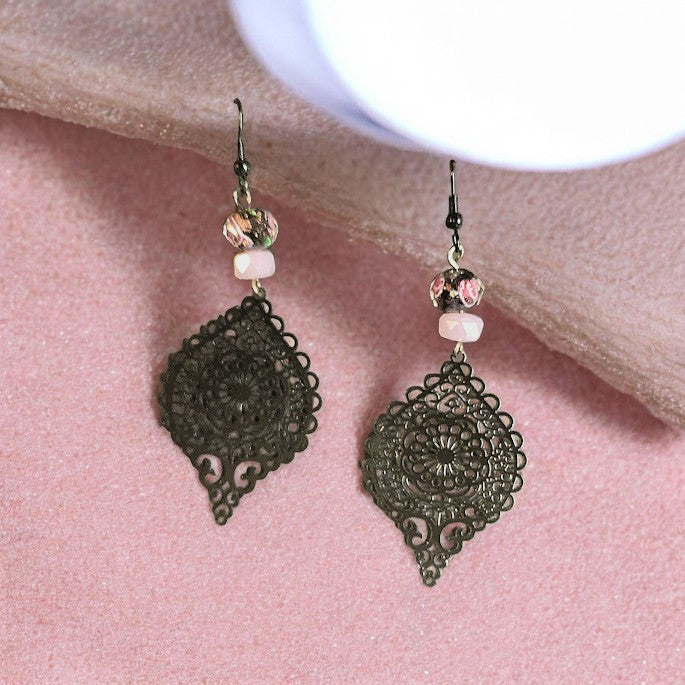 A pair of dangling earrings with ornate dark filigree teardrop charms, pink faceted beads, and clear crystal accents, displayed on a textured pink surface.