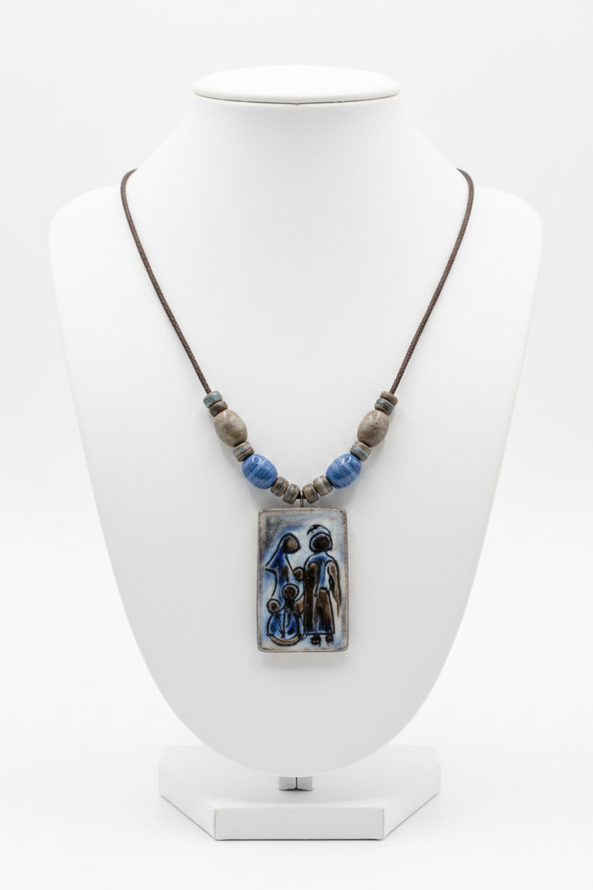 A ceramic pendant necklace displayed on a white mannequin bust. The necklace features a brown cord, blue and gray decorative beads, and a rectangular ceramic pendant depicting a stylized scene of figures in blue and brown tones.