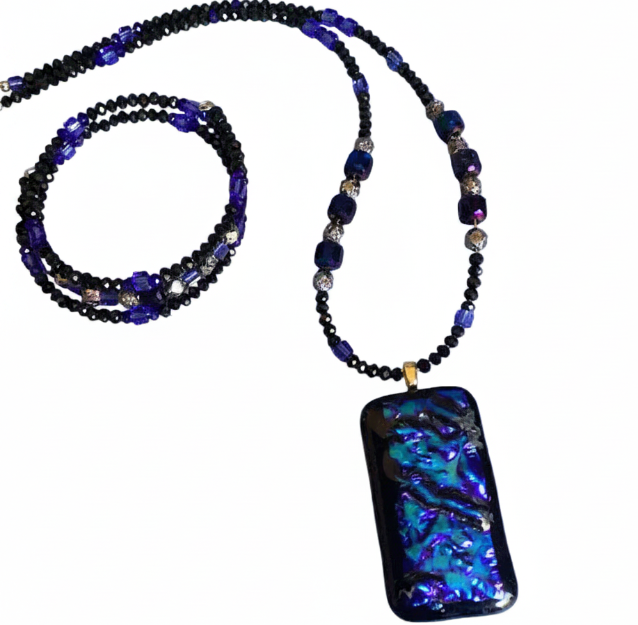 A beaded necklace featuring a large rectangular pendant with a black background and vibrant blue, purple, and green iridescent opal inlay. The pendant is suspended from a beaded chain composed of black beads, dark blue beads, and small silver-toned beads.