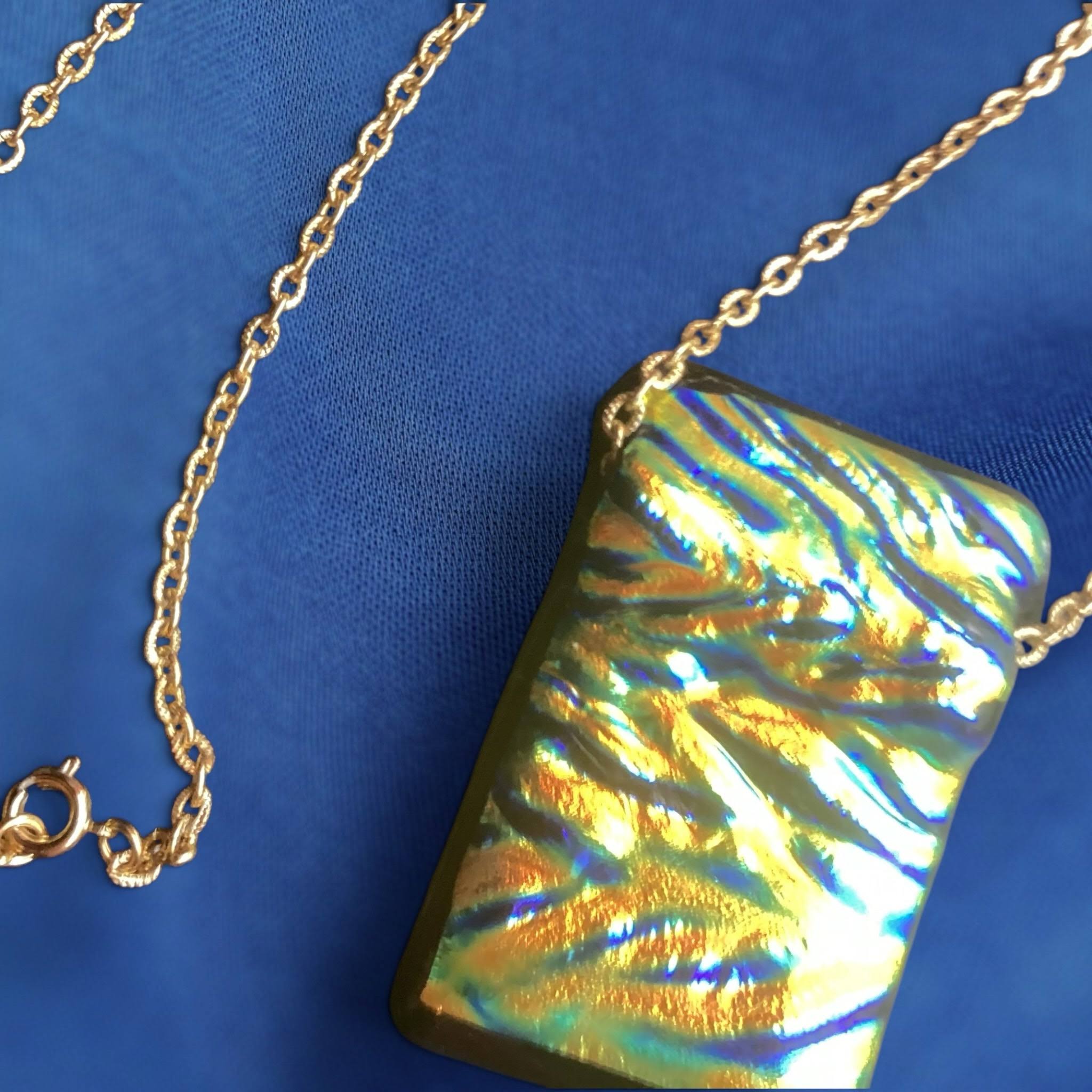 rectangular, iridescent dichroic glass pendant with a wavy, textured surface in shimmering shades of gold, green, and blue, suspended from a delicate, thin gold-colored chain with a small clasp