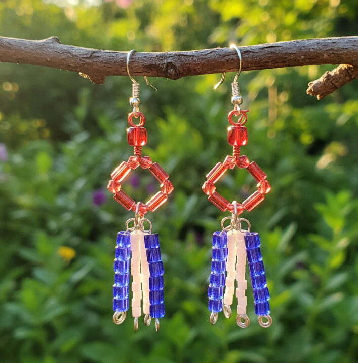 A pair of handmade beaded dangle earrings hanging from a tree branch outdoors. The earrings feature red geometric beads, blue and white beaded fringe, and silver fishhook wires, set against a blurred green garden background.