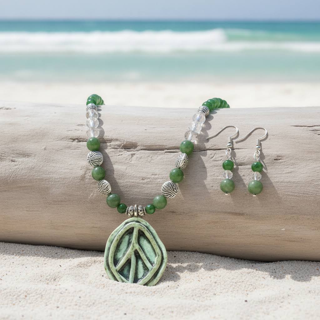 a handmade necklace and earring set featuring a large, oval-shaped, light green ceramic pendant with a carved peace sign, strung on a beaded chain of polished dark green jade and clear crystal beads, separated by ornate, textured silver metal beads, with matching drop earrings.