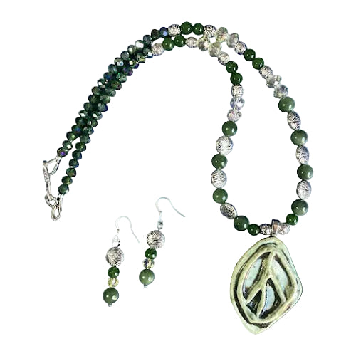a handmade necklace and earring set featuring a large, oval-shaped, light green ceramic pendant with a carved peace sign, strung on a beaded chain of polished dark green jade and clear crystal beads, separated by ornate, textured silver metal beads, with matching drop earrings.