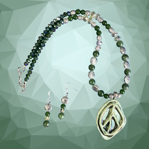 a handmade necklace and earring set featuring a large, oval-shaped, light green ceramic pendant with a carved peace sign, strung on a beaded chain of polished dark green jade and clear crystal beads, separated by ornate, textured silver metal beads, with matching drop earrings.