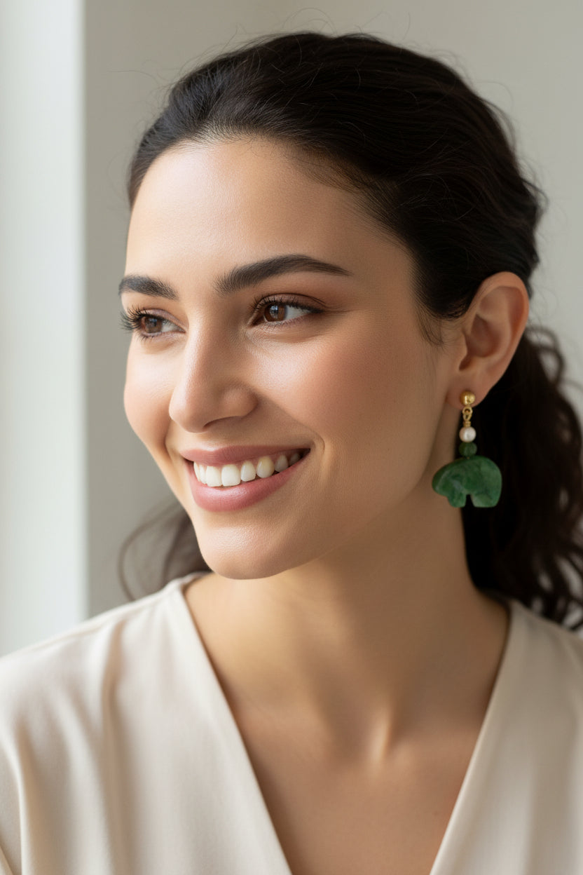 A pair of gold-tone drop earrings featuring carved green stone elephant charms, accented with a small green bead and a round white bead, displayed on draped cream satin fabric.