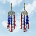 A pair of dangling earrings with ornate gold-tone filigree diamond-shaped tops and three hanging strands of faceted beads in royal blue, red, and clear, shown against a dark blue starry sky background.