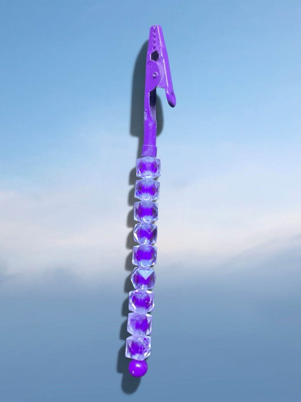 Heartfelt Creations purple metal roach clip featuring faceted crystal-like beads against a blue sky background.