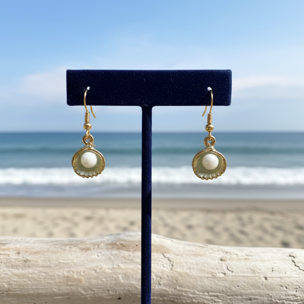 A pair of gold-tone dangle earrings with white pearl-like centers displayed on a black T-stand, photographed against a blurred beach and ocean background.