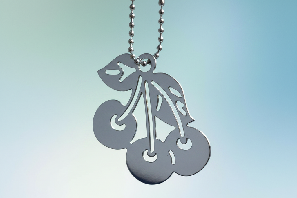 A cherry-shaped, silver plated over brass, keychain is hanging from a silver ball chain against a soft blue gradient background.